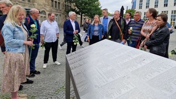 Auf dem Bild ist ein Teil der Gruppe mit weißen Rosen in der Hand zu sehen. Sie stehen hinter einer der zwei Gedenkstelen und lassen sich von Dr. Jochen Rath (dem Mann im dunklen Anzug in der Bildmitte), dem Leiter des Stadtarchivs Bielefeld, die Hintergründe zu dem Gedenkort erklären.