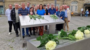 Auf dem Bild ist die Gruppe hinter einer von zwei Gedenkstelen vor dem Hauptbahnhof in Bielefeld zu sehen. In der Mitte ist die Regierungspräsidentin Anna Katharina Bölling mit blauem Kleid zu erkennen. Zur Erinnerung an die deportierten Menschen, die auf den Stelen aufgeführt sind, hat die Gruppe zuvor weiße Rosen darauf niedergelegt.