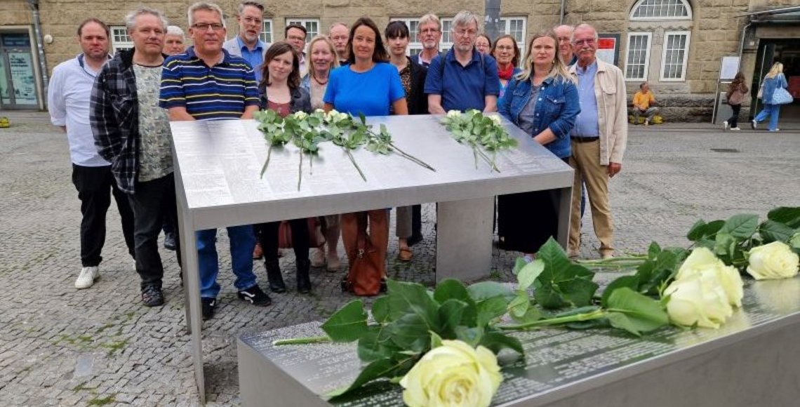 Auf dem Bild ist die Gruppe hinter einer von zwei Gedenkstelen vor dem Hauptbahnhof in Bielefeld zu sehen. In der Mitte ist die Regierungspräsidentin Anna Katharina Bölling mit blauem Kleid zu erkennen. Zur Erinnerung an die deportierten Menschen, die auf den Stelen aufgeführt sind, hat die Gruppe zuvor weiße Rosen darauf niedergelegt.